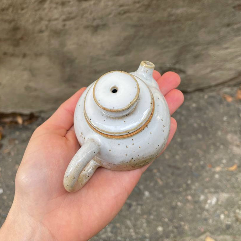 Hand holding a small ceramic teapot against a concrete wall.